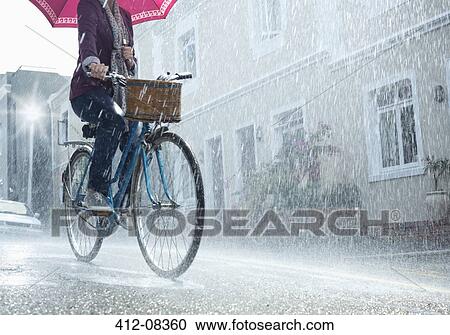 Stock Image - Woman riding bicycle with umbrella in rainy street. Fotosearch
