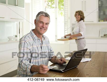 Stock Photo - Portrait of smiling senior man using laptop in kitchen. Fotosearch
