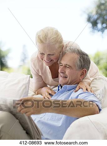 Stock Image - Senior couple reading newspaper on patio sofa. Fotosearch