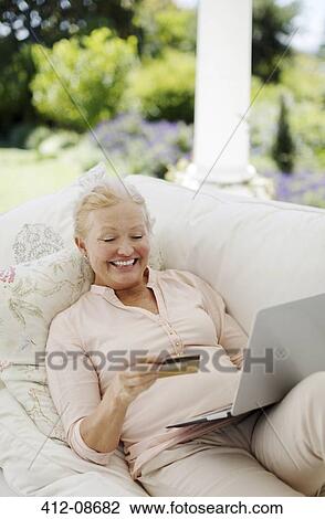 Stock Image - Senior woman shopping online on patio sofa. Fotosearch
