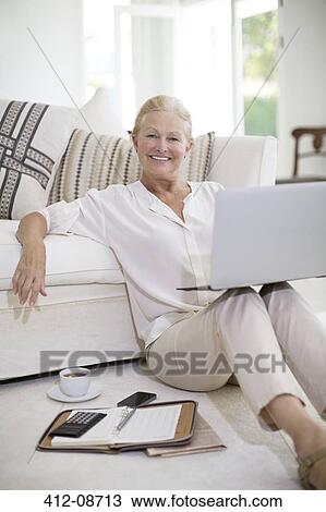 Stock Image - Senior woman using laptop on living room floor. Fotosearch