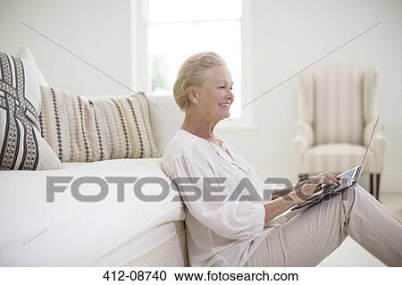 Stock Image - Senior woman using laptop on living room floor. Fotosearch