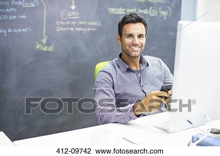 Stock Image - Businessman using cell phone at desk in office. Fotosearch