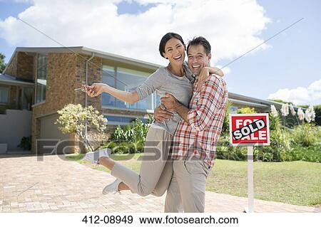 Stock Photo - Portrait of enthusiastic couple hugging outside house with For Sale sign. Fotosearch