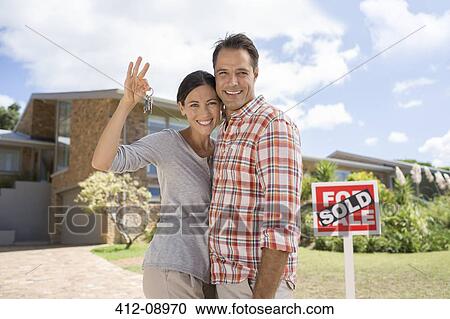 Stock Image - Portrait of smiling couple holding keys in front of new house. Fotosearch