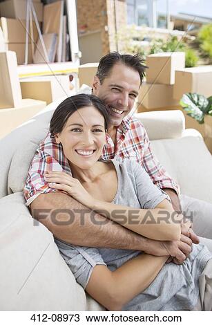 Stock Image - Portrait of smiling couple hugging on sofa in driveway near moving van. Fotosearch