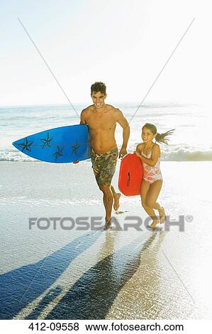 Stock Photo - Father and daughter carrying surfboard and bodyboard on beach. Fotosearch