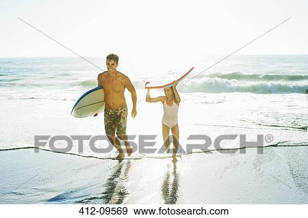 Stock Photo - Father and daughter carrying surfboard and bodyboard on beach. Fotosearch