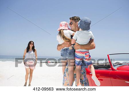 Stock Image - Father holding children on beach near convertible. Fotosearch