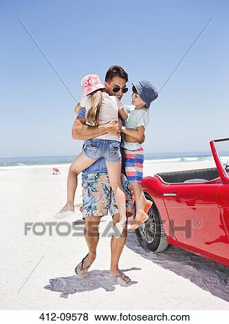 Stock Photo - Father hugging children on beach next to convertible. Fotosearch