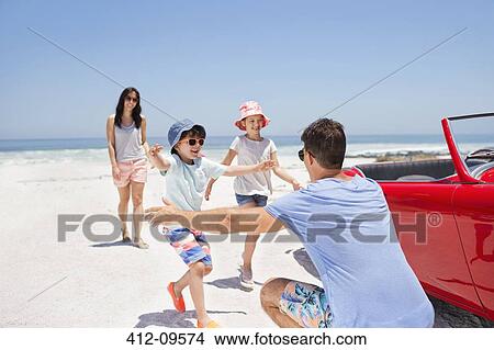 Picture - Father reaching to hug children on beach next to convertible. Fotosearch