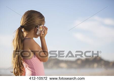 Stock Image - Girl listening to seashell on beach. Fotosearch