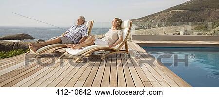Stock Image - Older couple relaxing in lawn chairs by pool. Fotosearch