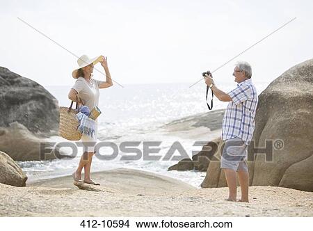 Picture - Older man taking pictures of girlfriend on beach. Fotosearch