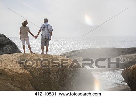 Stock Image - Senior couple holding hands on rocks at beach. Fotosearch