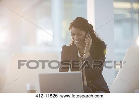 Stock Photograph - Businesswoman using laptop and cell phone in office. Fotosearch