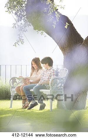 Brother and sister using digital tablet on bench under tree View Large Photo Image Picture - Brother and sister using digital tablet on bench under tree. Fotosearch