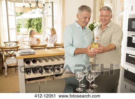 Stock Image - Senior me looking at wine bottle in kitchen. Fotosearch