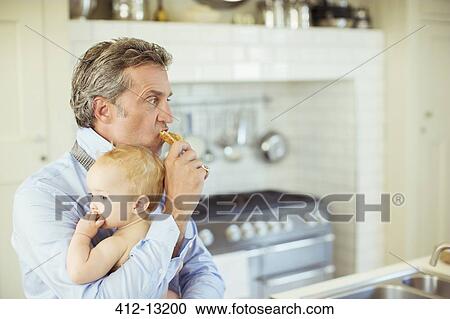 Stock Image - Father holding baby and eating breakfast. Fotosearch