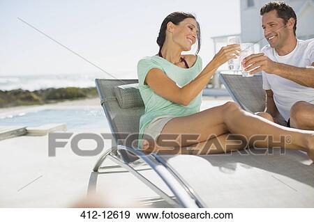 Stock Photo - Couple toasting water glasses in lounge chairs at poolside. Fotosearch