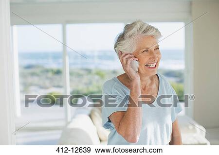 Stock Photo - Senior woman talking on cell phone in sunny living room. Fotosearch
