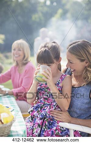 Stock Image - Girl drinking juice on mother's lap in backyard. Fotosearch