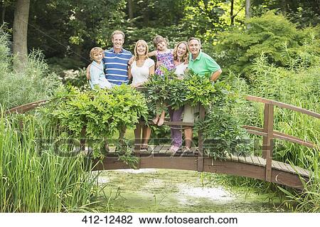 Stock Image - Multi-generation family standing on wooden footbridge over pond. Fotosearch