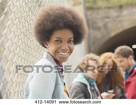 Stock Image - Woman smiling by chain link fence with friends in background. Fotosearch