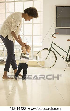 Stock Image - Mother helping baby boy walk in living room. Fotosearch