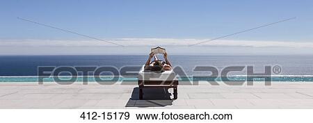 Stock Photo - Woman relaxing on lounge chair at poolside overlooking ocean. Fotosearch