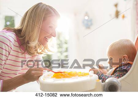 Stock Image - Mother feeding baby girl in high chair. Fotosearch
