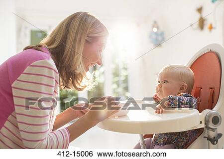 Stock Photograph - Mother feeding baby girl in high chair. Fotosearch