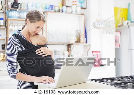 Stock Image - Mother with baby boy using laptop in kitchen. Fotosearch