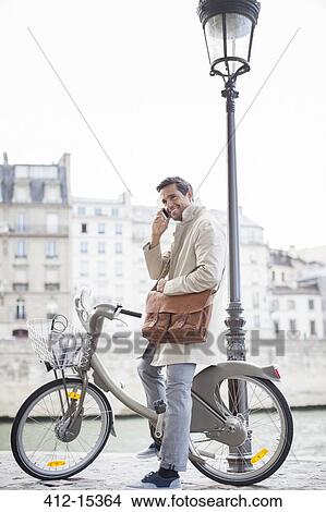 Picture - Businessman talking on cell phone on bicycle along Seine River, Paris, France. Fotosearch