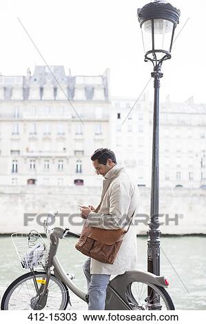 Stock Image - Businessman using cell phone on bicycle along Seine River, Paris, France. Fotosearch