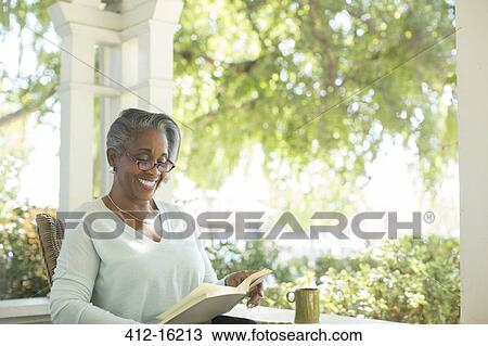 Stock Image - Happy senior woman reading book on porch. Fotosearch