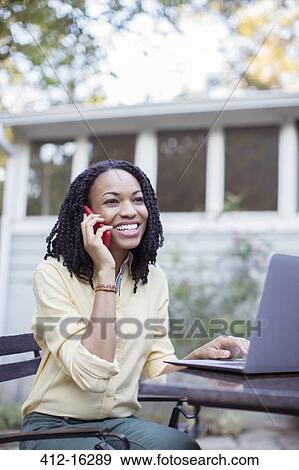 Stock Photo - Happy woman talking on cell phone at laptop on patio. Fotosearch