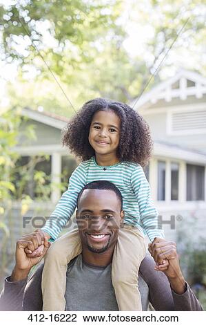 Stock Image - Portrait of smiling father carrying daughter on shoulders. Fotosearch