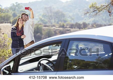Stock Photo - Couple taking self-portrait with cell phone outside car. Fotosearch