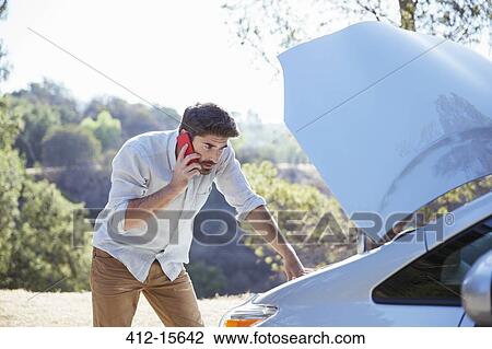 Stock Image - Man talking on cell phone and checking car engine at roadside. Fotosearch