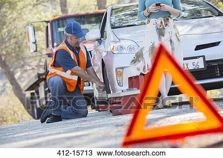 Stock Image - Roadside mechanic changing tire behind warning triangle. Fotosearch