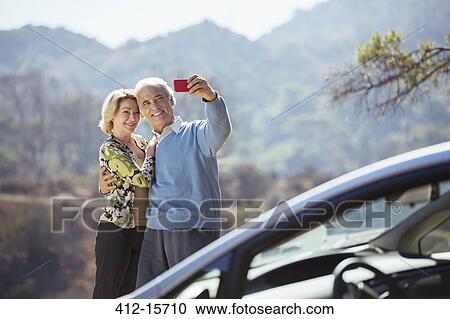 Stock Image - Senior couple taking self-portrait with cell phone outside car. Fotosearch