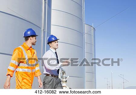 Stock Image - Businessman and worker walking near silage storage towers. Fotosearch