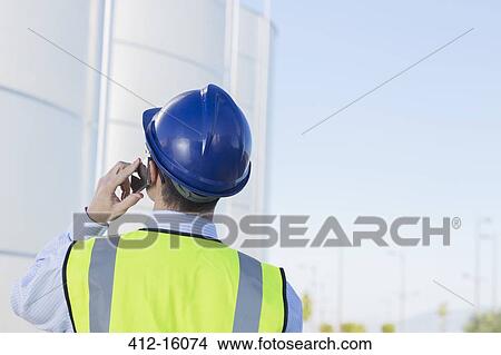 Picture - Worker talking on cell phone and looking up at silage storage towers. Fotosearch