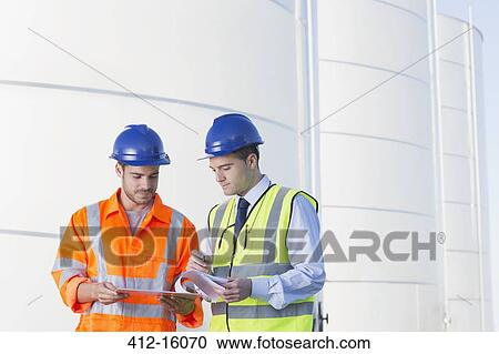 Stock Image - Workers reviewing paperwork next to silage storage towers. Fotosearch
