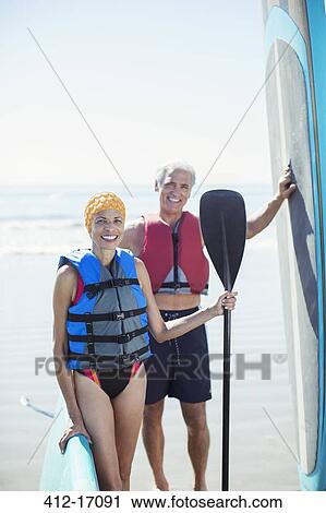 Stock Image - Portrait of senior couple with paddleboards on beach. Fotosearch