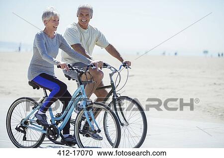 Stock Image - Senior couple riding bicycles on beach boardwalk. Fotosearch