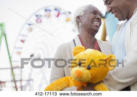 Stock Image - Senior couple with teddy bear hugging at amusement park. Fotosearch