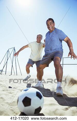 Stock Image - Senior men playing soccer on beach. Fotosearch