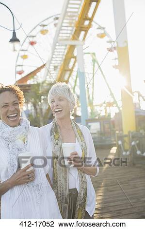 Stock Image - Senior women laughing and drinking coffee at amusement park. Fotosearch